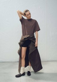 Pruhované tričko so širokými rukávmi v kombinácii s čiernymi šortkami. Dlhý hnedý vrchný odev prehodený cez plecia, čierne mokasíny a krémové ponožky. Hladká textúra.