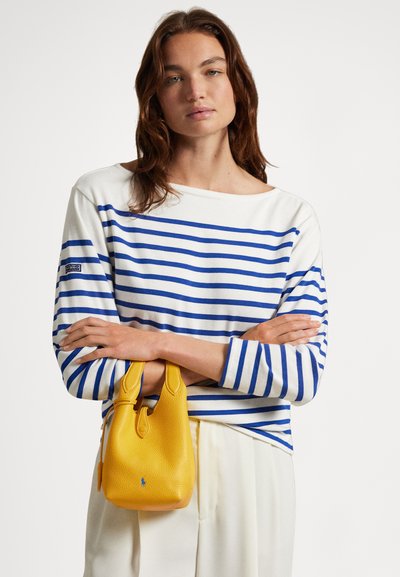 Bijeli top s plavim horizontalnim prugama, rukavi do pola podlaktice. Model drži teksturiranu žutu kožnu torbu s plavim detaljima logotipa.