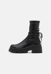 Black leather ankle boot with a rounded toe, thick treaded sole, elastic cuff, and decorative laces at the back. Smooth texture throughout.