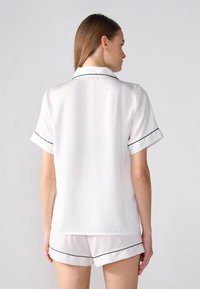 Valkoinen satiininen pyjama-setti mustilla reunuksilla. Lyhythihainen paita ja siihen sopivat shortsit, joissa on kaareva helma ja herkkä rakenne.