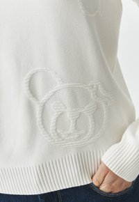 Suéter blanco de punto con un diseño texturizado de la cara de un oso, con dobladillos y puños acanalados, y un acabado suave. La mano en el bolsillo añade contexto.