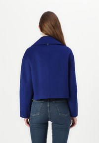 Afskåret blå uldjakke med spidse krave og en enkelt knap på ryggen. Kombineret med mørke denim jeans, set bagfra.
