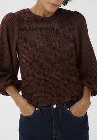 Blouse marron texturée avec smocks, manches bouffantes et ourlet volanté, associée à un jean en denim foncé.
