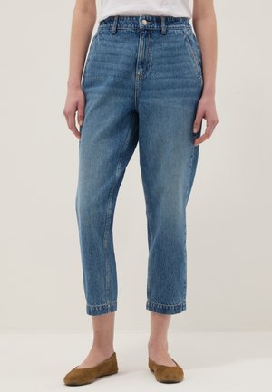 Persona che indossa jeans di denim blu a vita alta tagliati sopra le caviglie, abbinati a scarpe basse marroni e una camicia bianca, in piedi davanti a uno sfondo semplice.