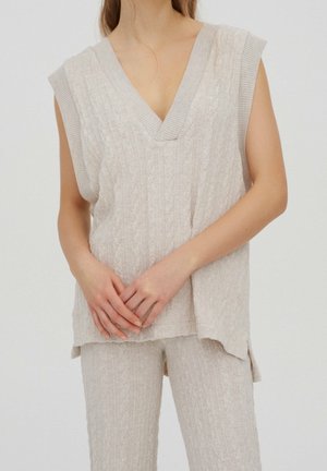 Femme portant un pull sans manches en maille torsadée beige à col en V et un pantalon texturé assorti, debout les mains jointes.