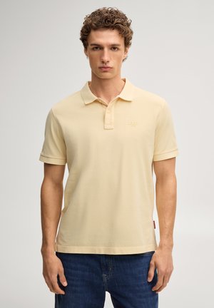 Młody mężczyzna z kręconymi włosami, ubrany w bladożółtą koszulkę polo i niebieskie dżinsy, stoi na tle gładkiego jasnego tła.