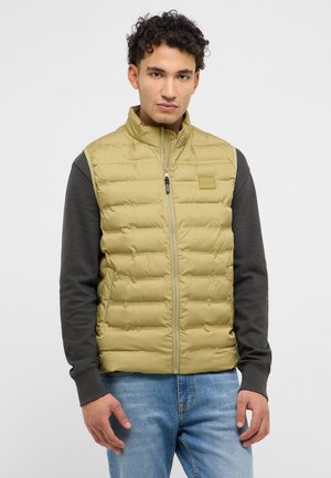 Junger Mann trägt eine hellolivfarbene, gesteppte ärmellose Weste über einem dunklen Langarmshirt und blauen Jeans, steht mit neutralem Gesichtsausdruck.