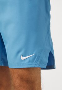 Zilā Nike šorti ar baltu logo kreisajā pusē, izgatavoti no gludas auduma ar sānu šķēlumiem mobilitātei.
