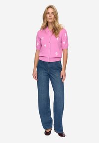 Růžový cardigan s krátkými rukávy, zdobený bílými vzory srdcí a listů, se zapínáním na knoflíky vpředu, doplněný o džíny s vysokým pasem v modré barvě a tmavou obuv.