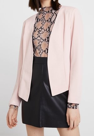 Femme portant un blazer rose clair sur un top imprimé serpent et une jupe en cuir noire, montrée des épaules jusqu'à mi-cuisse.