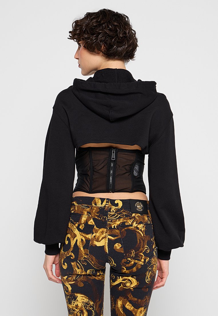 Sudadera negra corta con mangas largas y capucha, superpuesta a una blusa transparente estilo corsé con cremallera. Combinada con jeans multicolores de estampado.