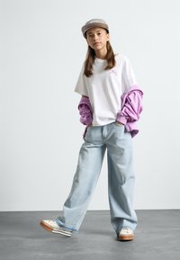 Maglietta bianca con logo rosa, sovrapposta a una felpa viola; jeans larghi azzurri chiari e sneakers beige con suole marroni. Cappellino grigio chiaro.