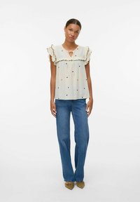 Haut beige clair à pois noirs et manches volantées, avec un décolleté en forme de trou de clé. Associé à un jean bleu à jambes larges et des ballerines marron clair.