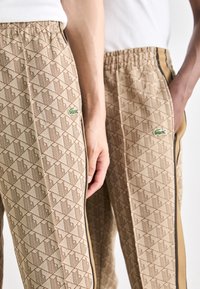 Beige raksti bikses ar ģeometrisku dizainu, ar elastīgu jostu, melnām sānu svītrām un mazu zaļu logotipa akcentu.