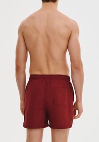 Bordowe kąpielówki wykonane z lekkiego materiału, z elastycznym paskiem w talii i jedną kieszenią z tyłu. Tekstura jest gładka i jednolita.