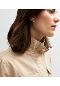 Beige jas met een hoge kraag, drukknopen en zijzakken. Model draagt zilveren ring oorbellen. Haar is schouderlang en golvend.