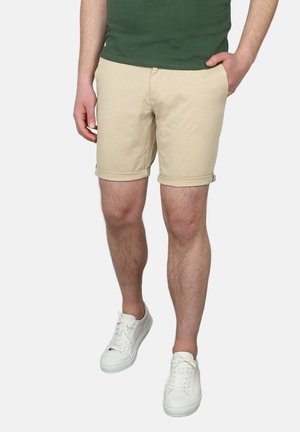 Man draagt beige opgerolde korte broek, witte sneakers en een groen overhemd, met één hand in de zak, staand tegen een effen achtergrond.