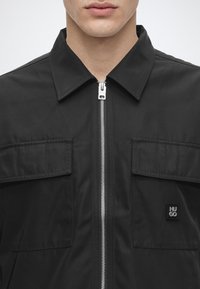 Homme portant une veste noire zippée avec un col et deux poches à rabat poitrine, dont une avec un petit patch logo Hugo.