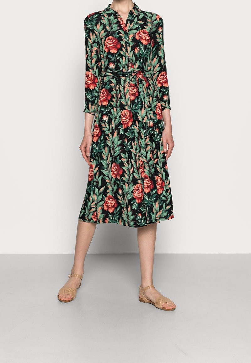 Donna che indossa un abito midi nero con motivi floreali rossi e foglie verdi, maniche lunghe, parte anteriore con bottoni, cintura in vita e sandali beige con punta aperta.