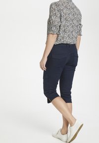 Marinblå capribyxor med flera fickor, rak skärning och slitsar på sidorna i fållen, kombinerat med en blommönstrad skjorta och vita sneakers.