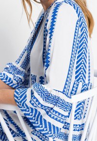 Tissu blanc avec des motifs géométriques bleus, doté de longues manches avec des poignets élastiques. Le design ouvert à l'avant améliore la respirabilité et le style.