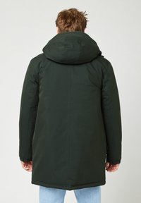 Man met een lange donkergroene winterjas met capuchon en lichtblauwe jeans, staand met de rug naar de camera tegen een effen lichte achtergrond.