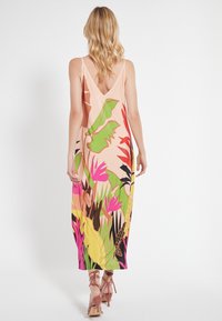Robe maxi sans manches rose clair avec un dos en V profond, arborant des motifs floraux tropicaux vibrants en vert, rose et noir. Texture lisse.