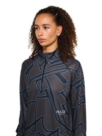 Kobieta o kręconych włosach, ubrana w czarno-szarą bluzkę z długim rękawem, z suwakiem do ćwiartki i logo "HALO" na klatce piersiowej.