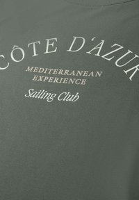 Grijze stof met witte en beige tekst met de woorden "Côte d'Azur Mediterranean Experience Sailing Club" in gebogen en rechte lijnen.