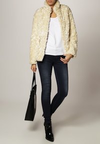 Cremefarvet falsk pelsjakke med høj krave, parret med en hvid top og mørkeblå jeans. En sort håndtaske og ankelstøvler fuldender looket.