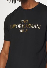 Svart bomull t-shirt med ett guldmetalliskt logotryck som inkluderar "EVA EMPORIO ARMANI MILAN," med rundad hals och korta ärmar.