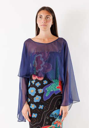 Donna dai lunghi capelli scuri indossa una blusa blu trasparente sopra un abito floreale senza maniche con motivi audaci e colorati, in piedi su sfondo bianco.