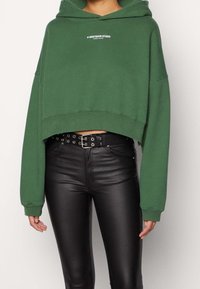 Persona che indossa un hoodie verde accorciato con testo bianco e pantaloni in finta pelle nera a vita alta con cintura a grommet.