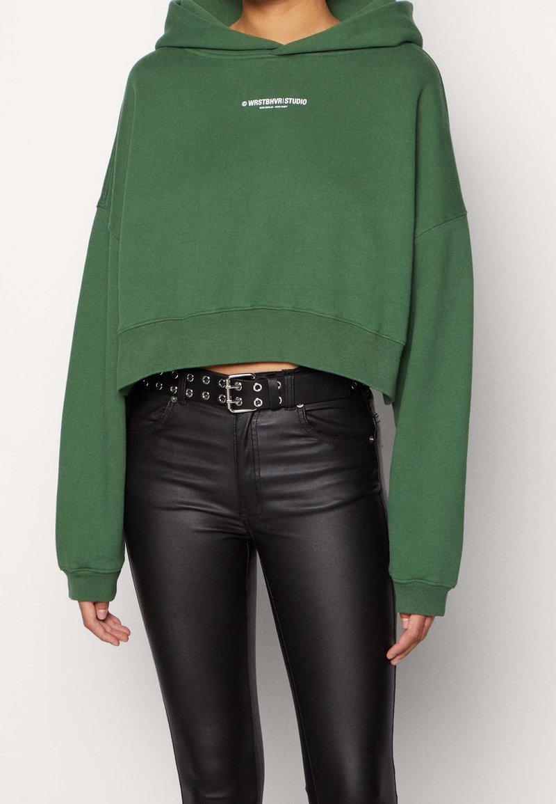 Persona che indossa un hoodie verde accorciato con testo bianco e pantaloni in finta pelle nera a vita alta con cintura a grommet.