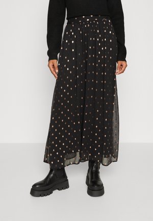 Jupe plissée noire avec un motif de pois dorés, fabriquée en tissu léger. Associée à des bottes noires à semelle épaisse.