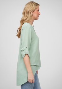 Top verde chiaro e testurizzato con maniche tre quarti, polsini a risvolto e orlo curvo, abbinato a jeans blu.