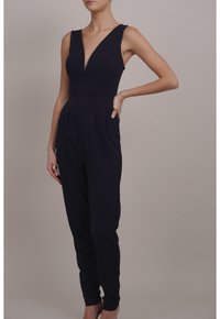 Mujer vestida con un mono negro sin mangas y un escote en V profundo, de pie con una mano en la cadera y la otra relajada a su lado.