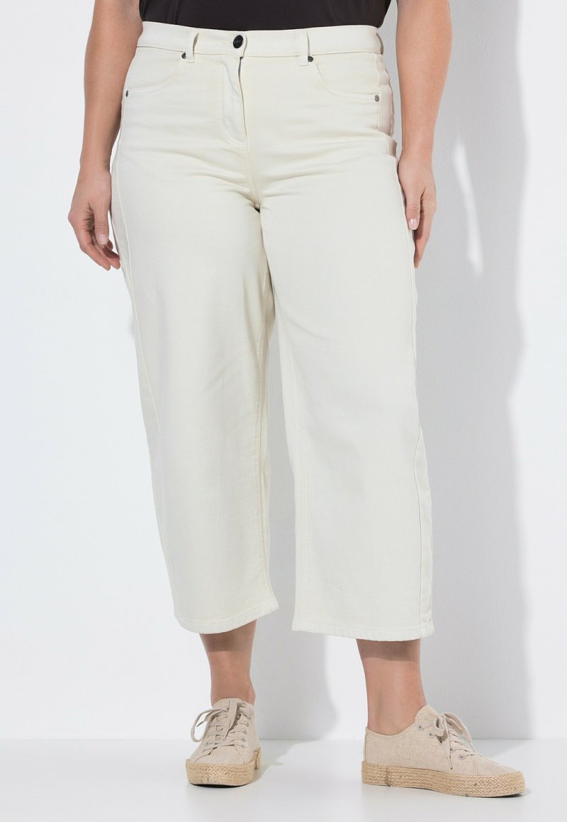 Kobieta ubrana w kremowe, szerokie, krótkie spodnie, w zestawieniu z jasnobeżowymi sneakersami na sznurowadła, stojąca na białym tle.