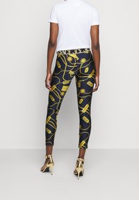 Svarta leggings med ett guldkedjemönster och olika etiketter. Midjebandet visar "REA" i fet, kontrasterande text.