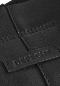 Svart läderprodukt med synliga sömmar och en strukturerad yta. En rektangulär läderlapp med präglad text "DEPECHE".