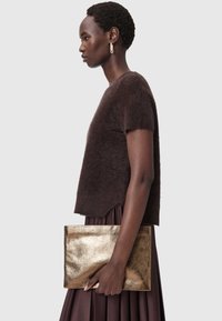 Brun stickad kortärmad tröja med luddig yta kombinerad med en plisserad brun kjol. Modellen håller en glänsande gyllene metallickuvertväska.