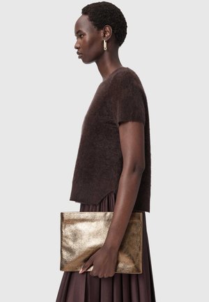 Brauner, flauschiger Strickpullover mit kurzen Ärmeln, kombiniert mit einem braunen, plissierten Rock. Das Model hält eine glänzende Clutch aus goldfarbenem Metall.