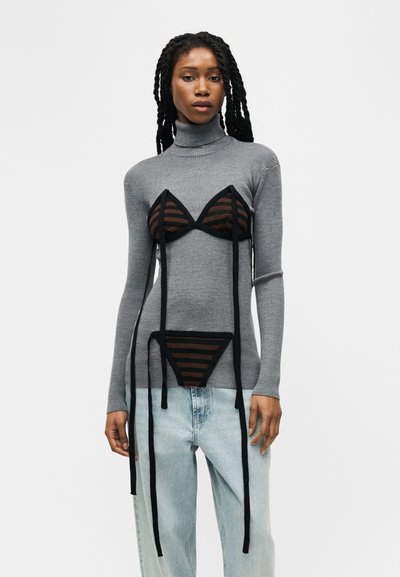 Femme aux longs cheveux tressés portant un pull à col roulé gris superposé à un ensemble de lingerie en maille noire rayée et un jean bleu clair.