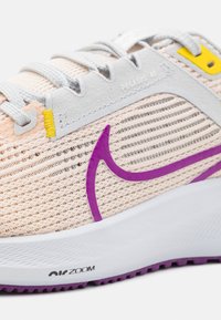 Gros plan d'une chaussure de course beige Nike Pegasus 40 avec swoosh violet, lacets gris, accents jaunes et semelle blanche marquée Air Zoom.