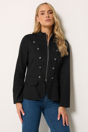 Femme aux longs cheveux blonds porte une veste noire double boutonnage zippée avec des boutons argentés et un jean bleu, debout sur un fond blanc.