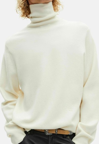 Suéter de cuello alto color crema hecho de tela suave, con mangas largas, corte relajado y un logo sutil. Combinado con un cinturón marrón y pantalones negros.