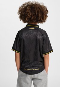 Svart kortärmad tröja med geometriskt mönster, gult krage och ärmdetaljer, med "INTERNATIONALS" och "hummel" märken på baksidan.