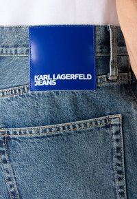 Detailní záběr na zadní kapsu modrých denimových džín s velkou modrou koženou nášivkou označenou "Karl Lagerfeld Jeans."