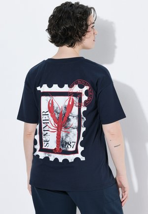 Personne aux cheveux courts et bouclés portant un t-shirt bleu marine avec un motif homard rouge et le texte « SUMMER 1987 » dans le dos.