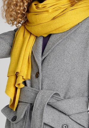 Kędzierzawe włosy i żółty szalik na osobie w szarym wełnianym płaszczu z paskiem i dużymi guzikami.